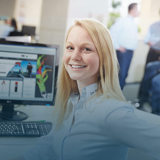 Junge Frau mit langen blonden Haaren vor einem Monitor, dreht sich in Richtung Kamera und lächelt freundlich.