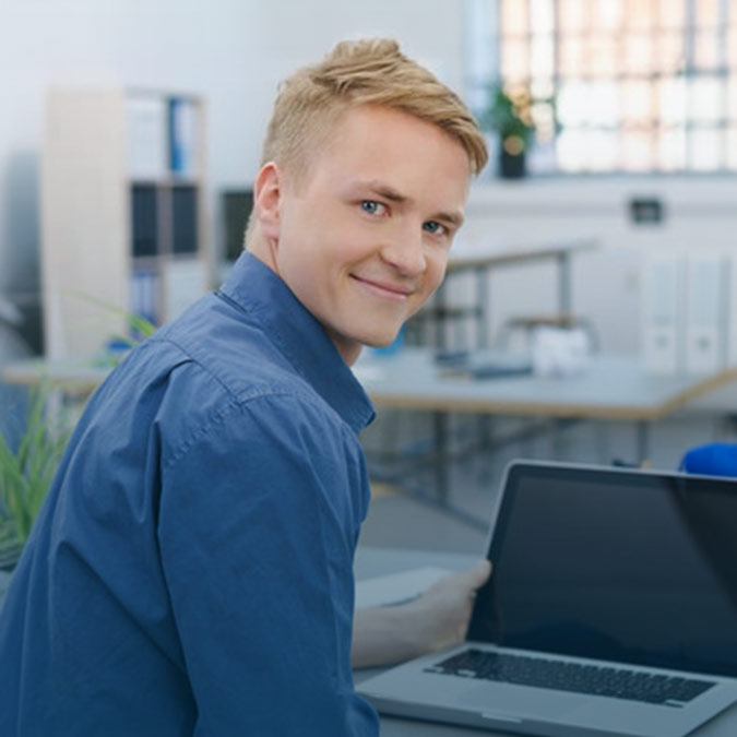 Junger Mann an einem Laptop, schaut über die Schulter und lächelt freundlich.