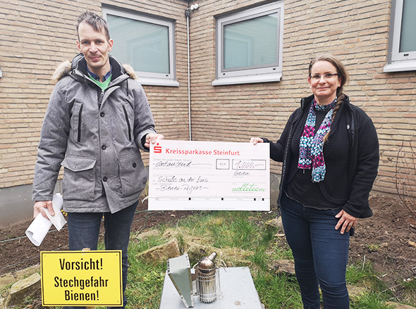 Zwei Personen rechts und links, dazwischen ein übergroßer Scheck, gelbes Schild "Vorsicht Stichgefahr: Bienen"